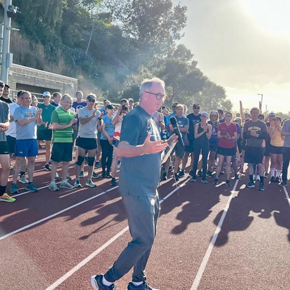 San Diego Track Club's Coach Paul Greer making a speech to a large group of athletes outside on a track.