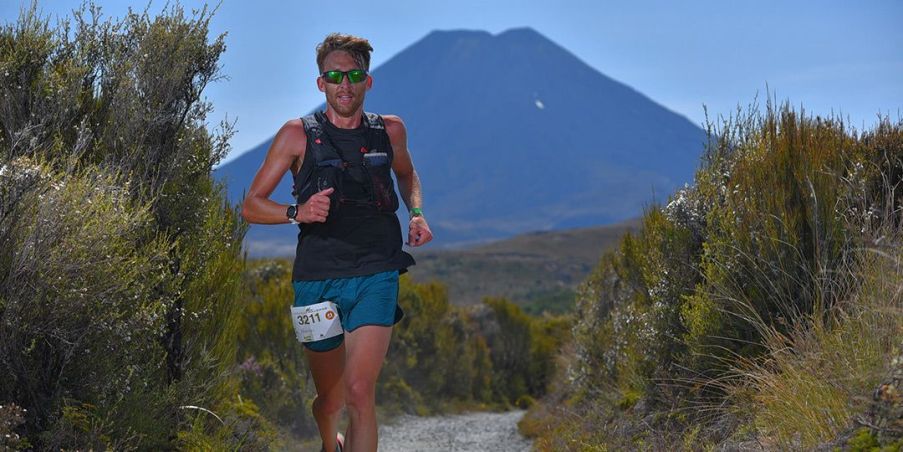 An athletic looking male wearing a black tank top and blue running shorts with a race bib attached, running on a trail surrounded by tall green shrubs with a large blue mountain in the distance behind him. 