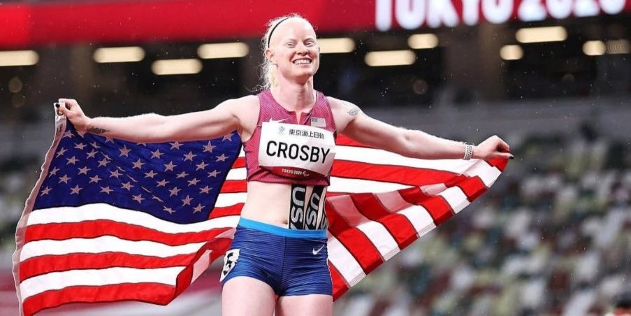 Team Injinji Athlete Kym Crosby wearing a USA National Track and Field team race kit and bib with her arms outstretched holding an American flag behind her signifying her bronze medal in the Paralympics.