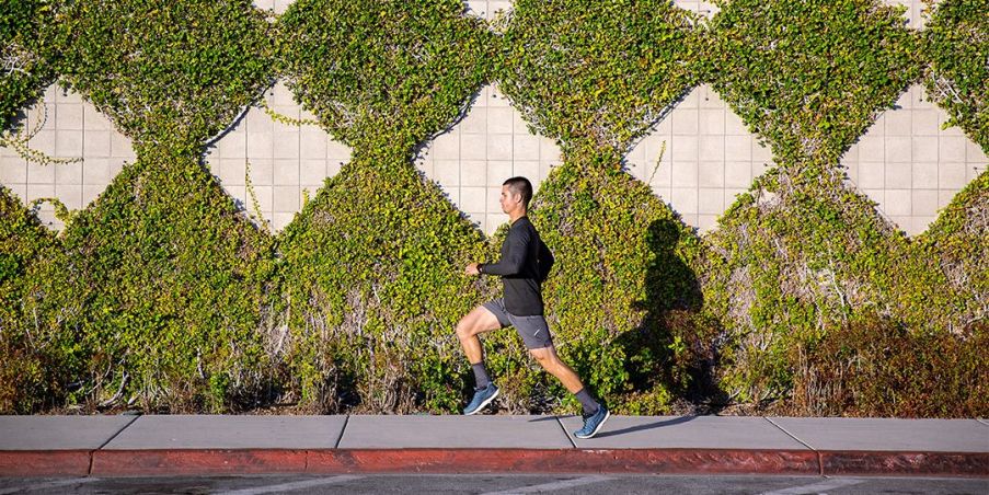 A man running on a sidewalk in front of a tall ivy covered wall. 