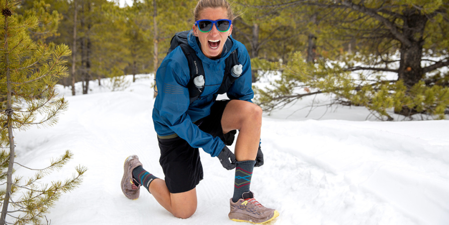 Ultra runner Courtney Dauwalter down on one knee adjusting her Injinji Trail Midweight Crew sock among snow and pine trees.