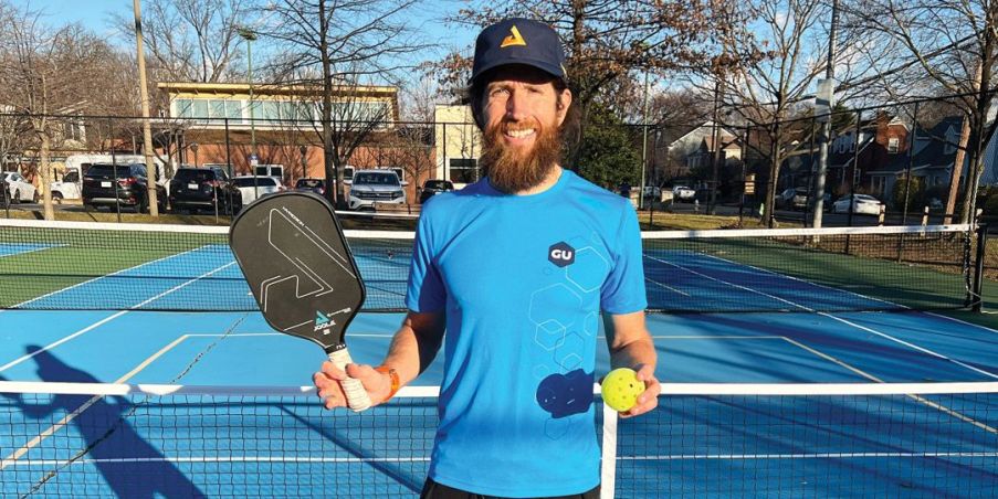 Mike Wardian with a pickleball paddle and ball standing on a pickleball court. 