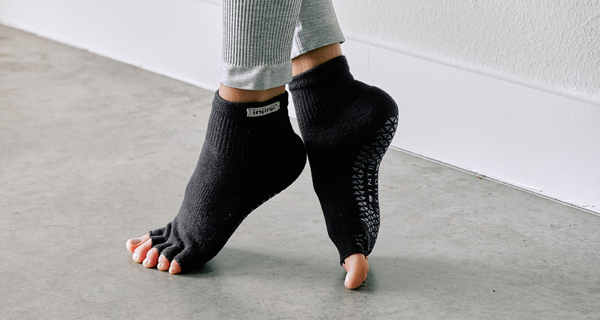 Close up of a woman standing with crisscrossed feet pointing her toes wearing Injinji X Pointe Studio Grip Toeless Mini-Crew in Black.