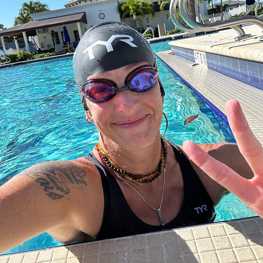 Karin Bachrodt wearing athletic swimming gear and giving a peace sign at the edge of a lane in a swimming pool.