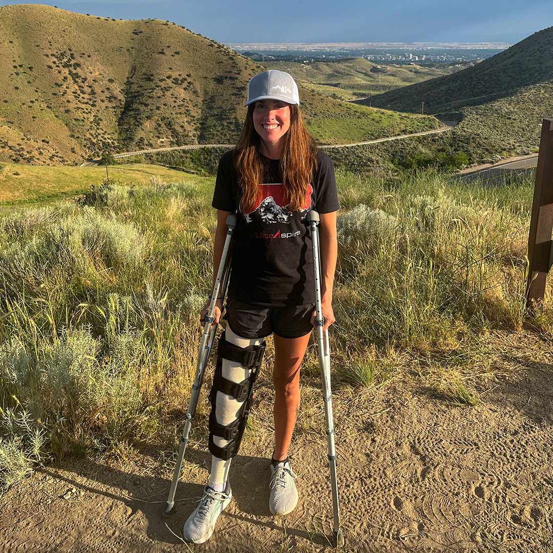 Trish Arends standing upright with crutches with a mountain trail behind her. 