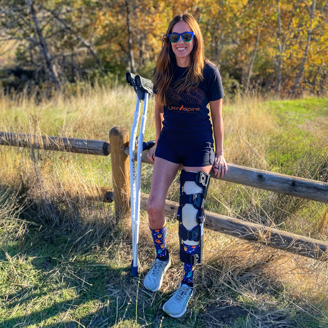 Trish Arends wearing a full leg stabalizing device while leaving on a wooden fence near a trail. 