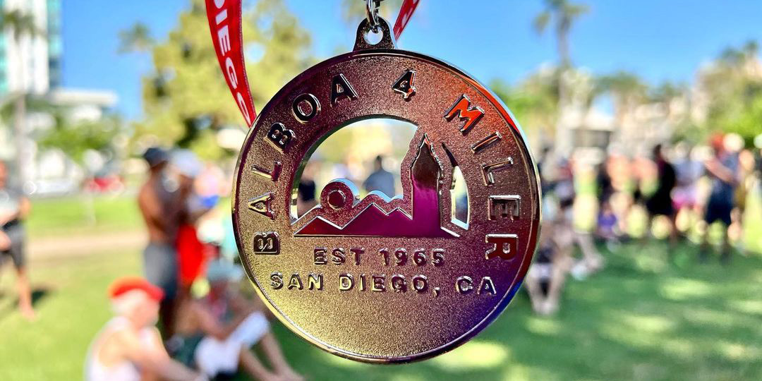 A closeup of a gold medal given to the winners of the Balboa 4 Miler race in San Diego, with people and green grass out of focus in the background. 