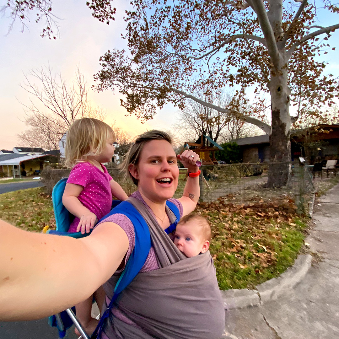 Cate Barrett on a walk carrying her newborn in a sling on her front side with her first child on her back. 