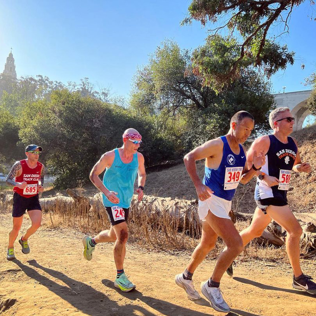 A group of four middle aged, males wearing multicolored shorts and tanktops, running on a dirt trail with downed trees in the background. 