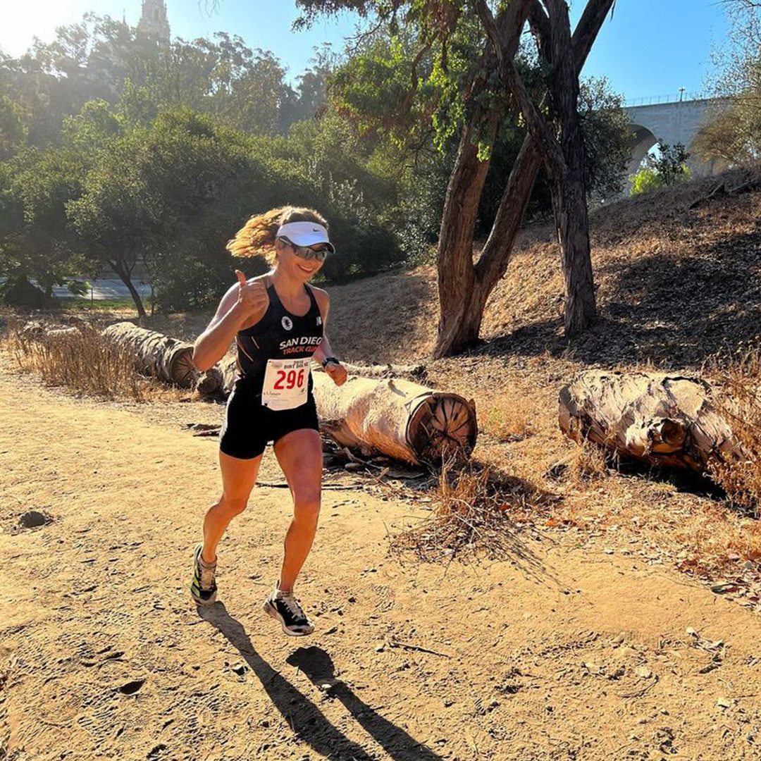 A female running on a dirt trail, wearing a black tank top and shorts, with downed trees in the background on a sunny day. 