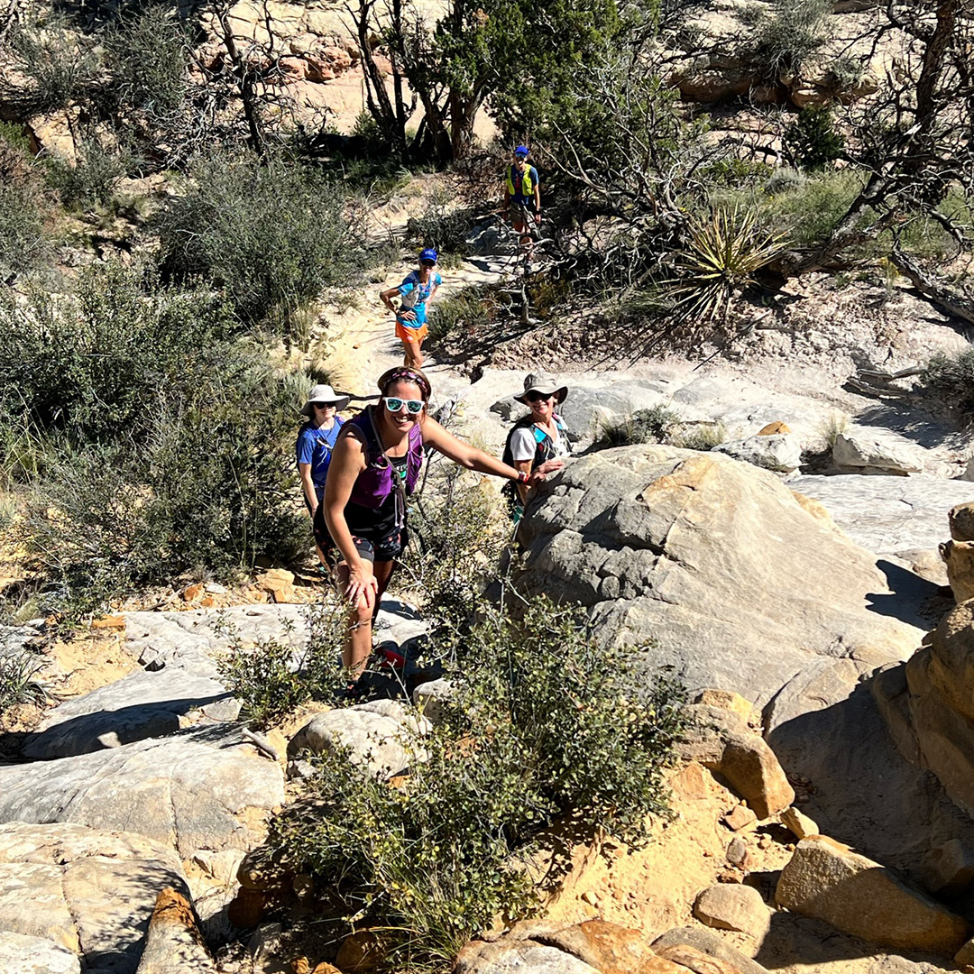 Looking down on a group of hikers from above. They're smiling and turned toward the camera, wearing sunglasses and sunhats in the dusty, high desert landscape.