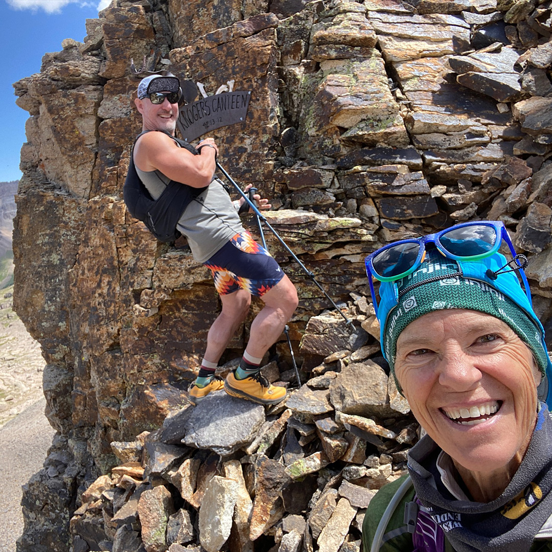 Tanned and happy Ultra runner, Meghan Canfield, smiles at the camera. Behind her is a male hiker standing on a pile of granite stones, smiling and pointing to a sign that says "Krogers Canteen".