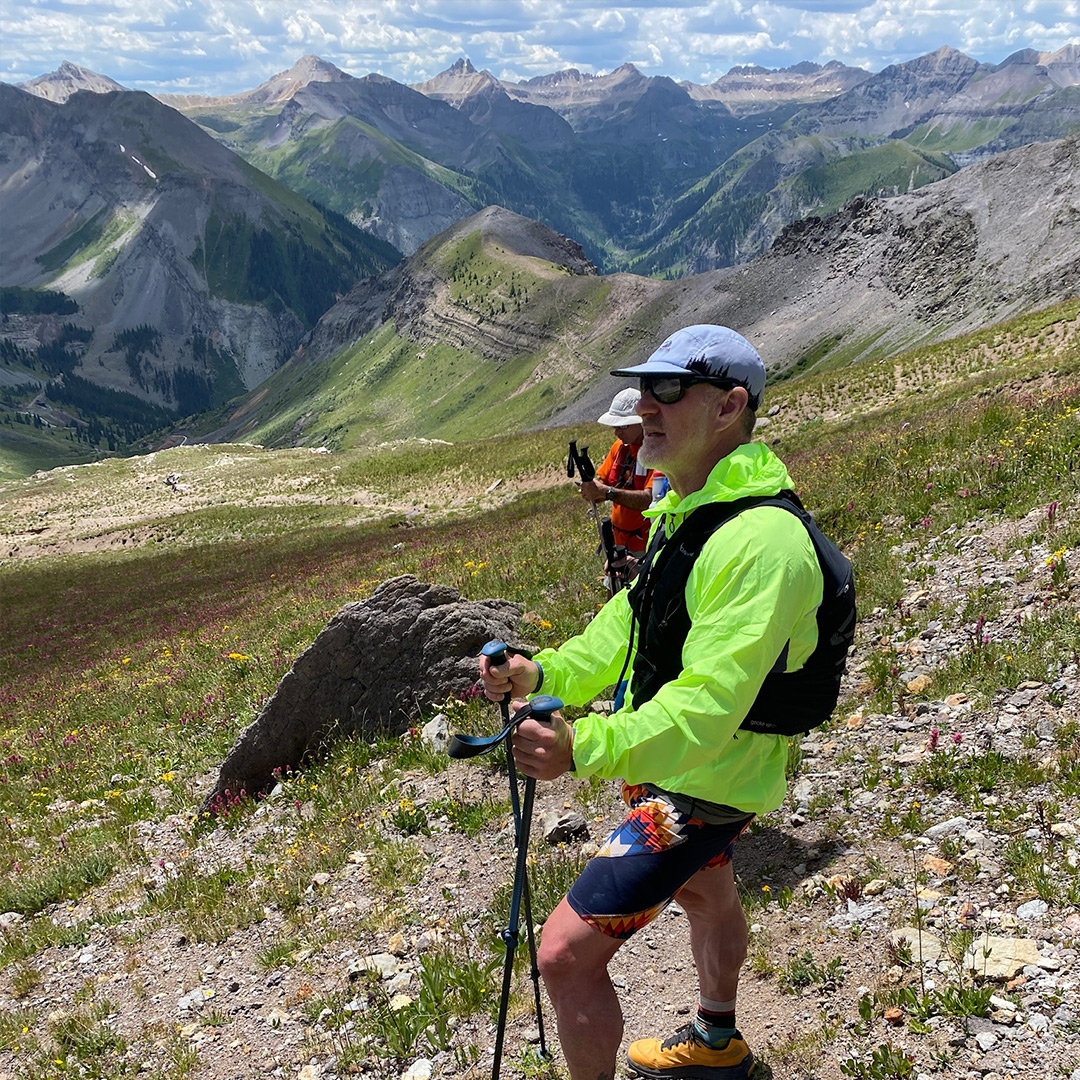 A older, white male hiker standing in his fluorescent green windbreaker, ball cap, running shorts and shoes, holding hiking poles in his hands. A wide view of rugged mountains is in the background.