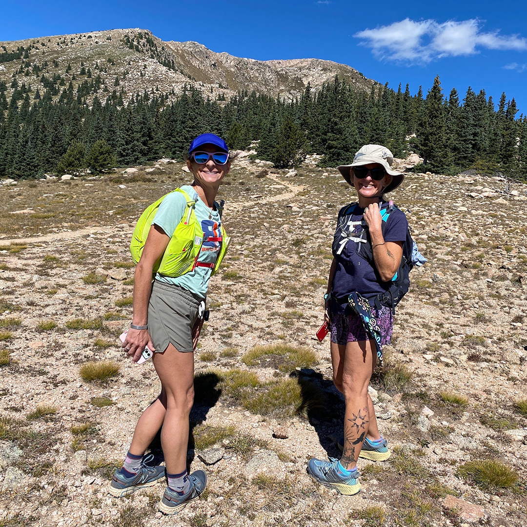 Two female hikers turned and smiling toward the camera among the high desert, alpine landscape of shrubbery and pine trees. 