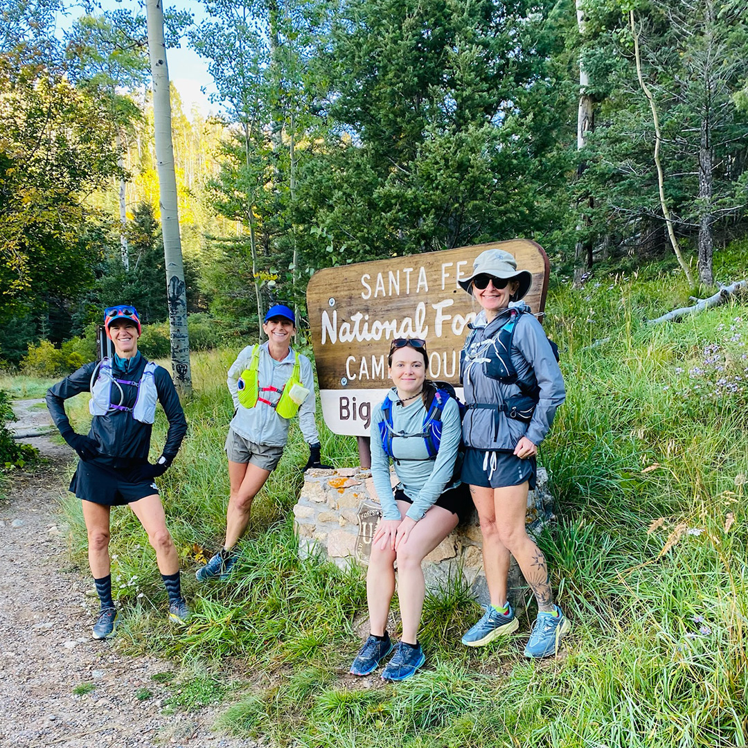 A group of female hikers smiling at the camera, posing next to the Sante Fe National Forest parks sign, among a grassy slope and varied green foliage in the background.