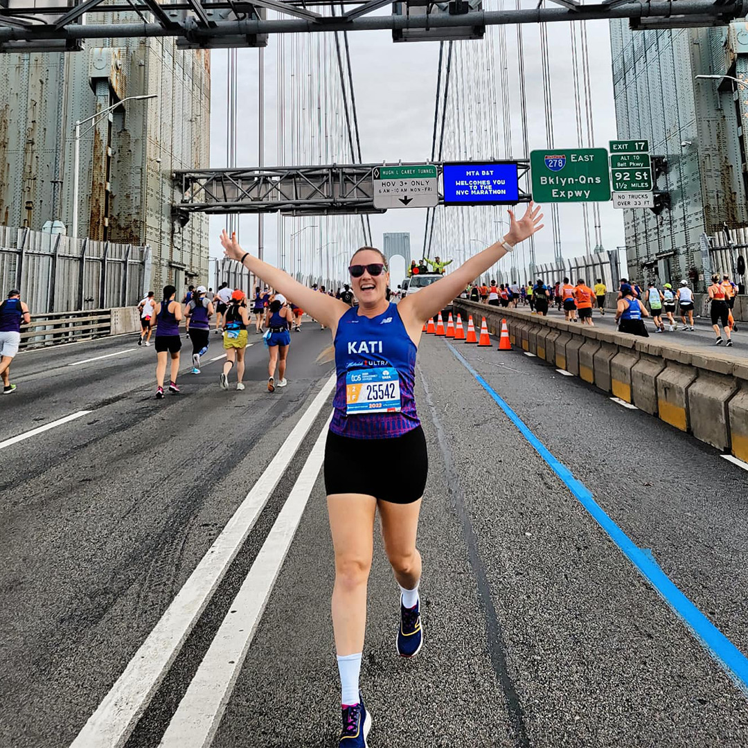 Kati Keenan excitedly running over a large city bridge during a marathon with her arms in the arm. 