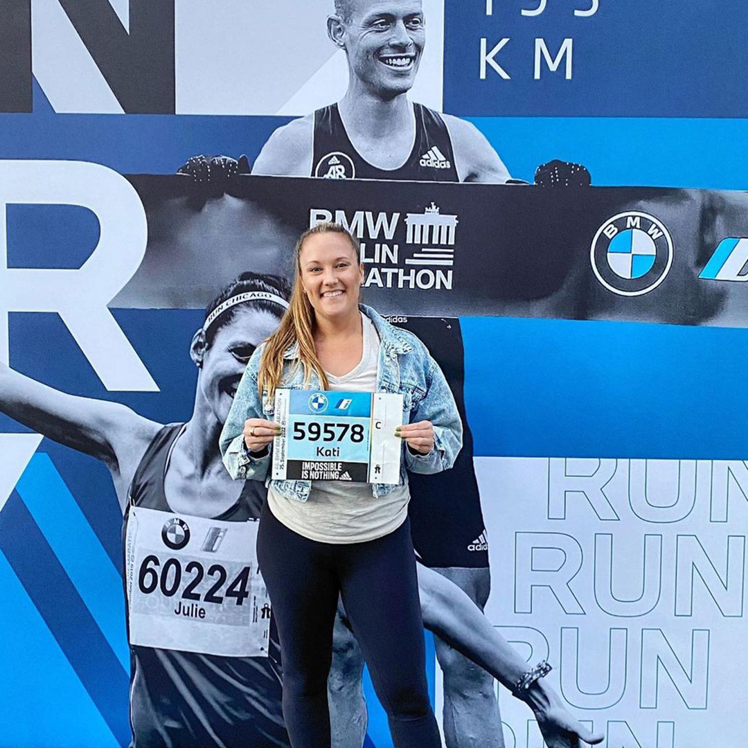 Marathon runner and Team Injinji athlete, Kati Keenan standing in front of a marathon race wall holding her race bib. 