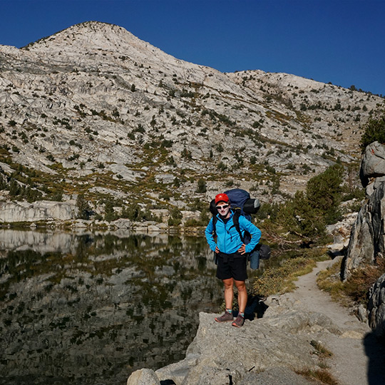 Male backpacker standing next to a lake with the reflection of the mountains on the water. 